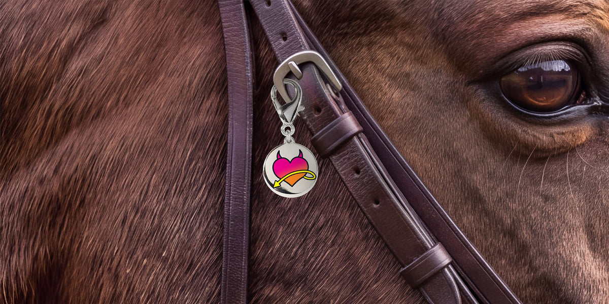 A close-up of a horse's face and bridle, featuring a round silver metal ID tag style charm with a pink heart with devil horns and yellow arrow tail across it design hanging from the buckle.