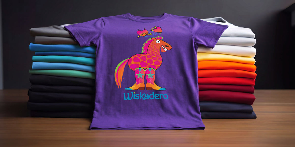A purple short-sleeve t-shirt with a colorful cartoon horse graphic and the word 'Wiskadero' printed below it, displayed in front of stacked folded shirts in multiple colors on a wooden surface.