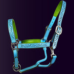 Full front view of the Wiskadero Halo and Horns padded leather halter. Highlights the turquoise blue leather with deeply tooled halo, horn, and heart motifs, vibrant lime green padded leather lining at the crown, noseband, and cheeks, and polished chrome hardware against a dark purple background.