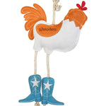 Close-up back view of the Wiskadero What the Cluck toy showing the white suede body with a stamped orange Wiskadero logo patch, orange tail feathers, natural rope pull-through legs, and turquoise suede cowboy boots with embroidered white stars.